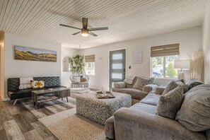 Ein modernes Wohnzimmer mit einem schwarzen Ledersofa, einem grauen Ottomanen und einem grauen Sofa. Es gibt einen Deckenventilator, ein Bild an der Wand und ein Fenster mit Blick ins Freie.