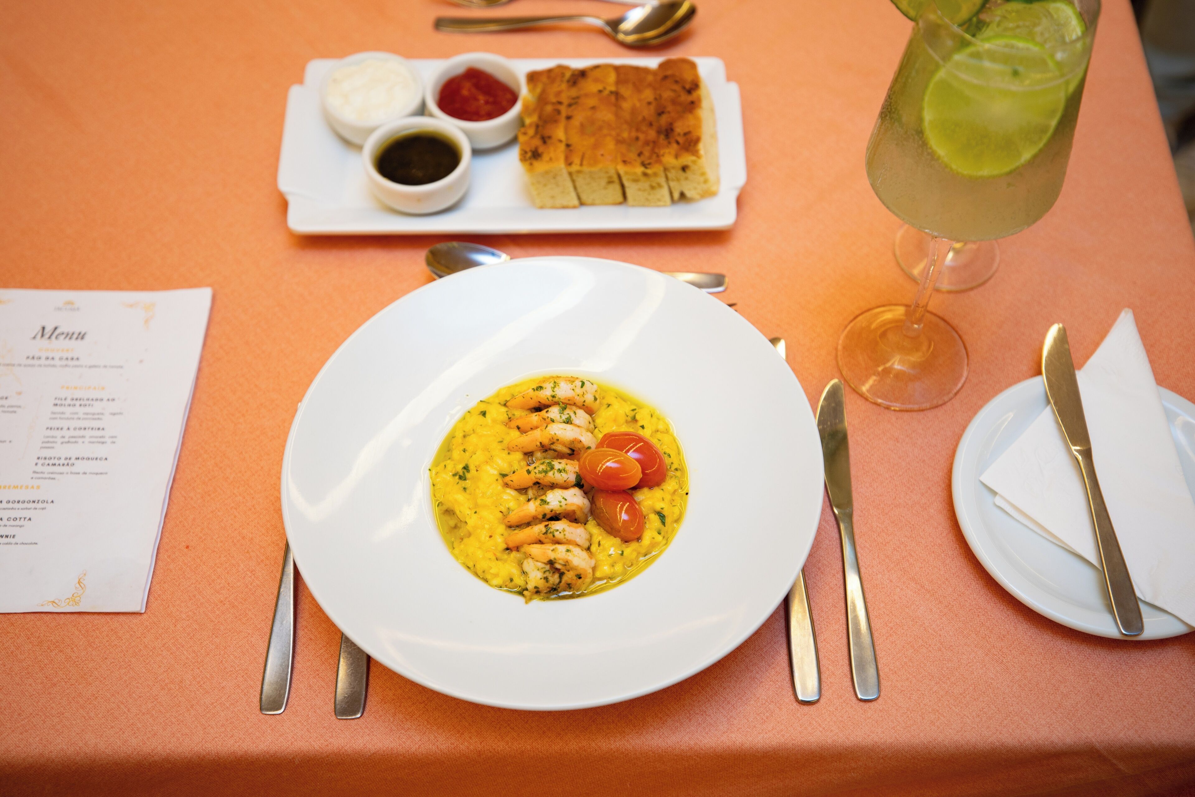 A table set with a plate of seafood risotto, a glass of lime drink, and a small plate of bread and condiments.