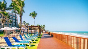 A wooden boardwalk with blue lounge chairs and umbrellas, palm trees, and a beach view.