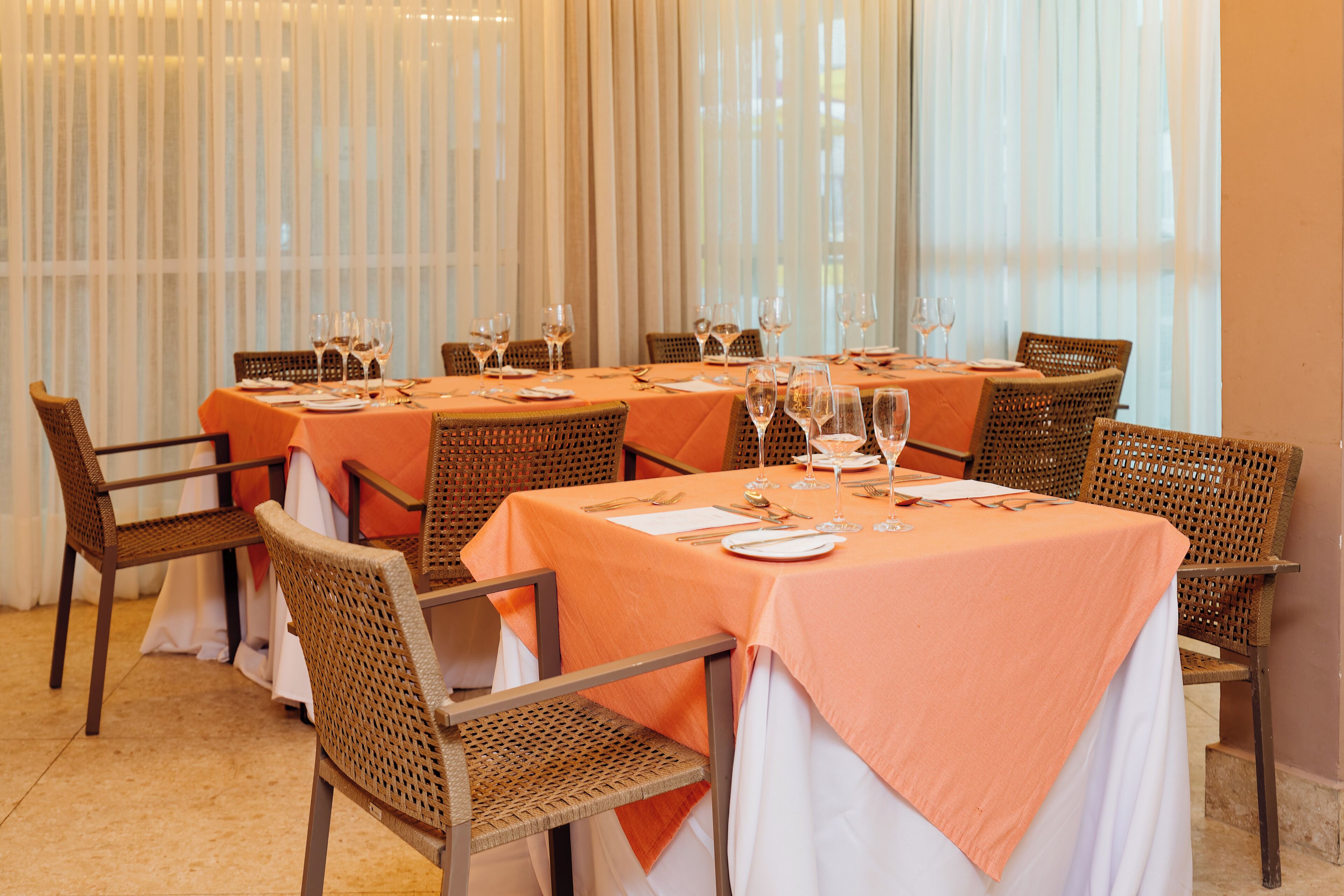 A dining area with tables set for six, each with a white tablecloth and orange napkins.