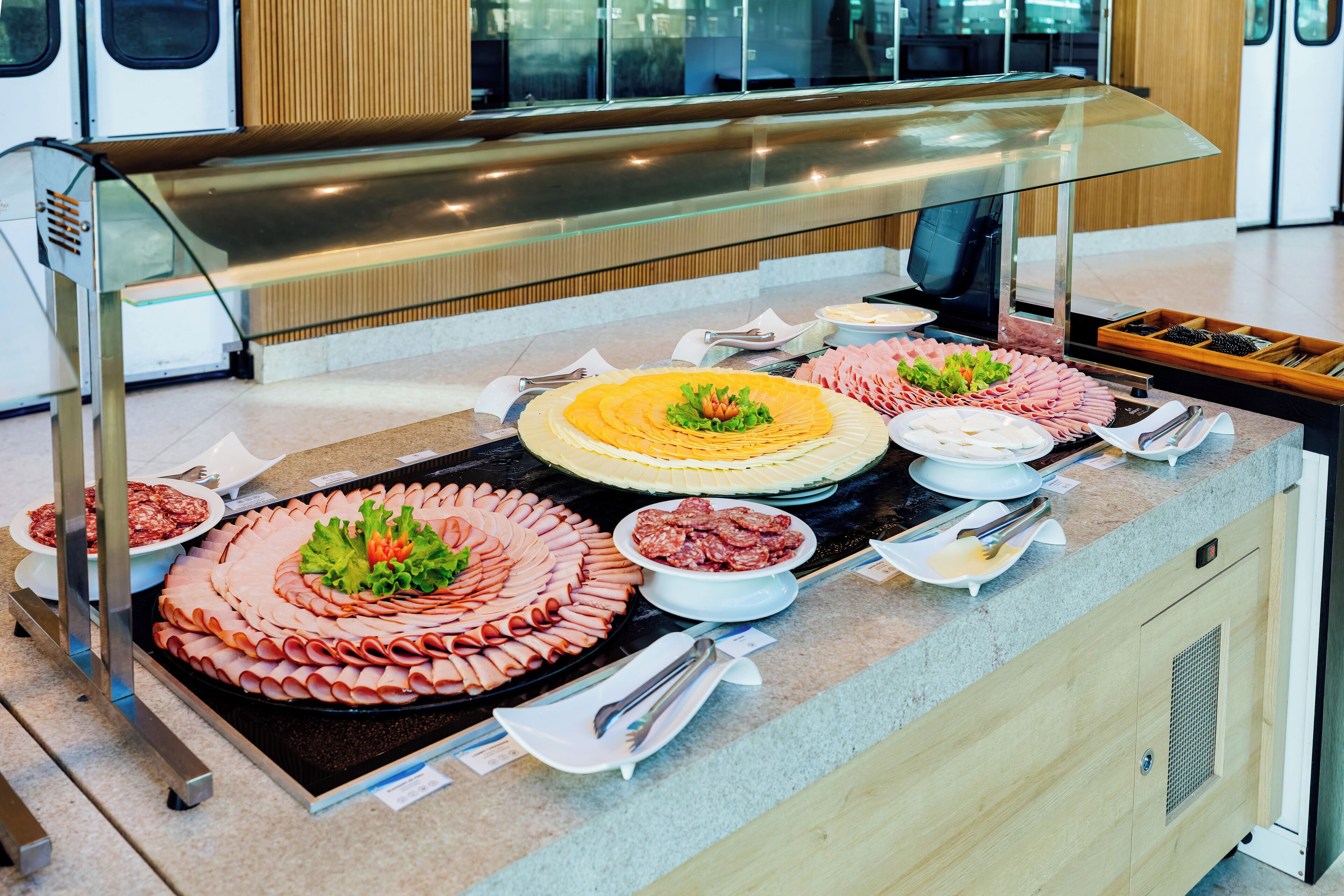 A buffet with various food items, including sliced meats and a large salad, arranged on a counter with a glass display case above.