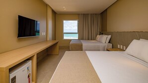 A hotel room with a bed, a TV, a window with a view of the sea, a desk, and a minibar.