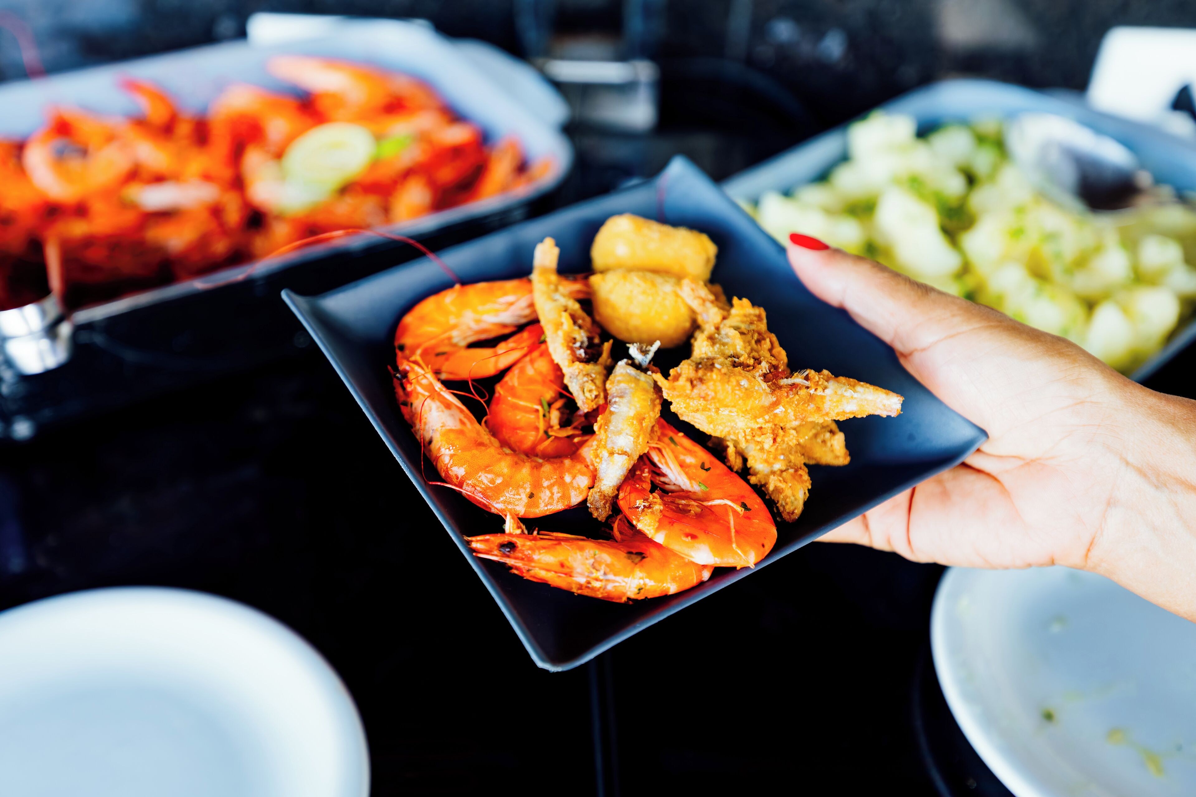 Uma mão segura um prato com camarões e alimentos fritos, com outros pratos ao fundo.