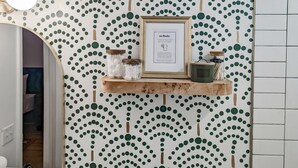 Un baño con papel tapiz verde y blanco a lunares, un estante de madera con frascos y un letrero enmarcado.