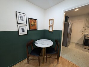 Una zona de menjador petita amb una taula rodona i quatre cadires, una cuina amb nevera i forn, i una porta que condueix a una altra habitació.