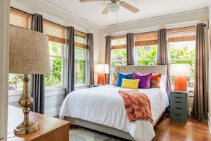 A bedroom with a bed, curtains, a ceiling fan, and a window with a view of greenery.