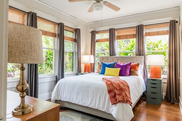A bedroom with a bed, curtains, a ceiling fan, and a window with a view of greenery.