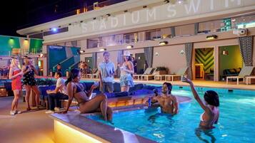 A swimming pool area with a large sign reading 'STADIUM SWIM' and people relaxing around the pool.