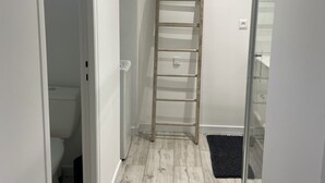 Kapea käytävä, jossa seinää vasten nojaa puinen tikapuu, valkoinen ovi, joka johtaa kylpyhuoneeseen, ja pieni musta matto lattialla.