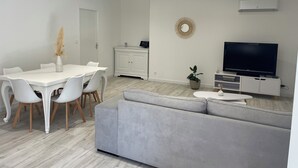 Moderni olohuone, jossa on harmaa sohva, ruokapöytä tuoleineen, televisio ja valkoinen kaappi.