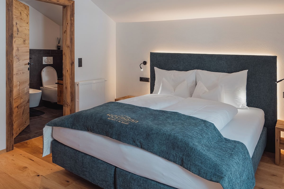 Une chambre avec un grand lit, un plancher en bois et une vue sur la salle de bain par une porte ouverte.