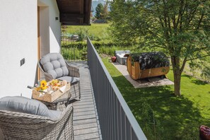 En balkon med fletningsmøbler, grill i træ og udsigt over en græsplæne med træer.