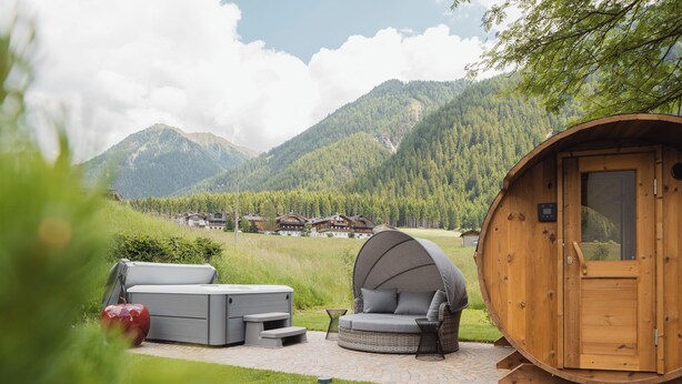 A wooden cabin with a covered seating area and a hot tub, surrounded by a lush garden and mountains in the background.