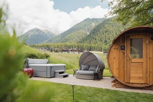 Uma cabana de madeira com área de estar coberta e banheira de hidromassagem, rodeada por um jardim exuberante e montanhas ao fundo.