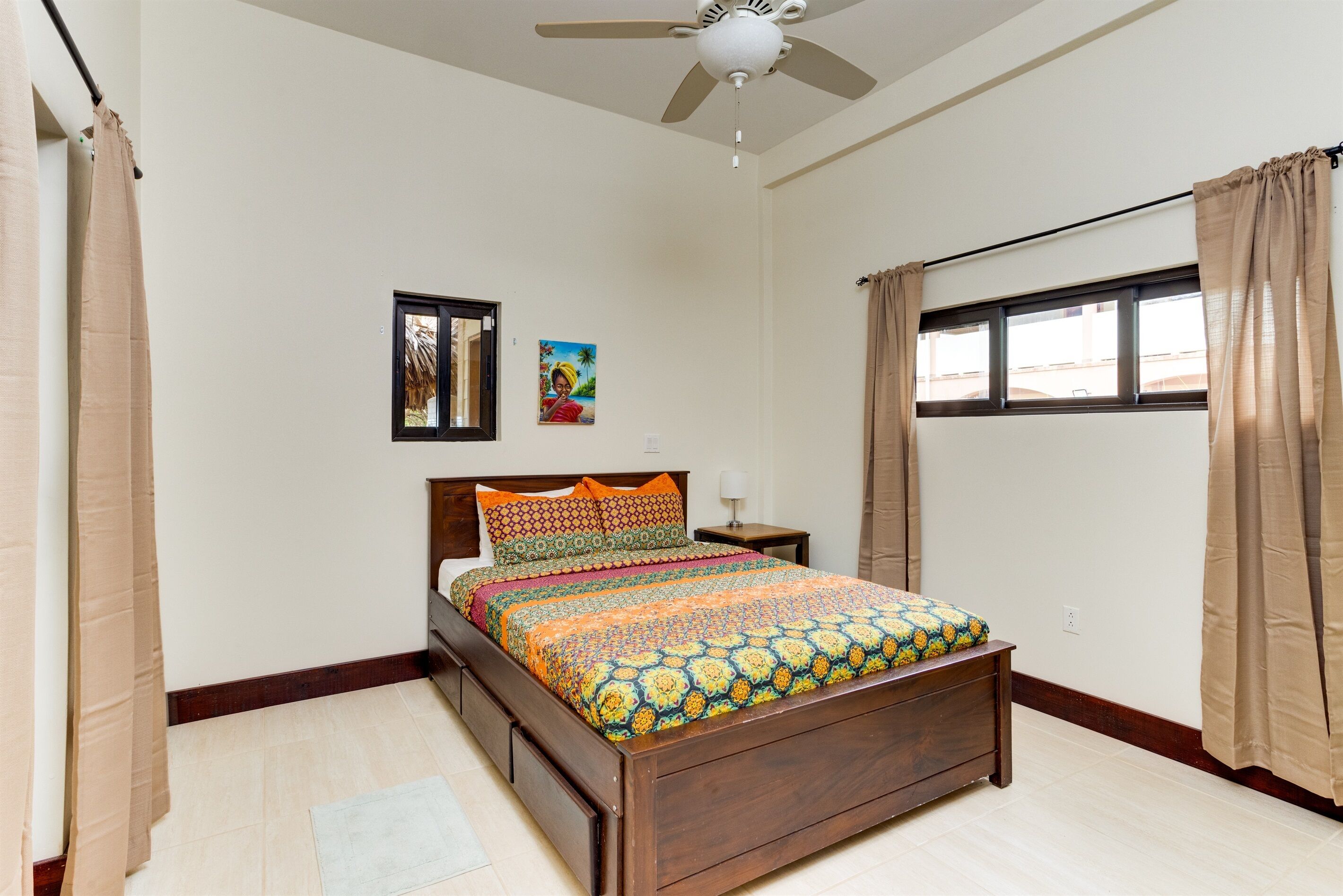A bedroom with a wooden bed, beige curtains, a ceiling fan, a window with curtains, and a small nightstand with a lamp.