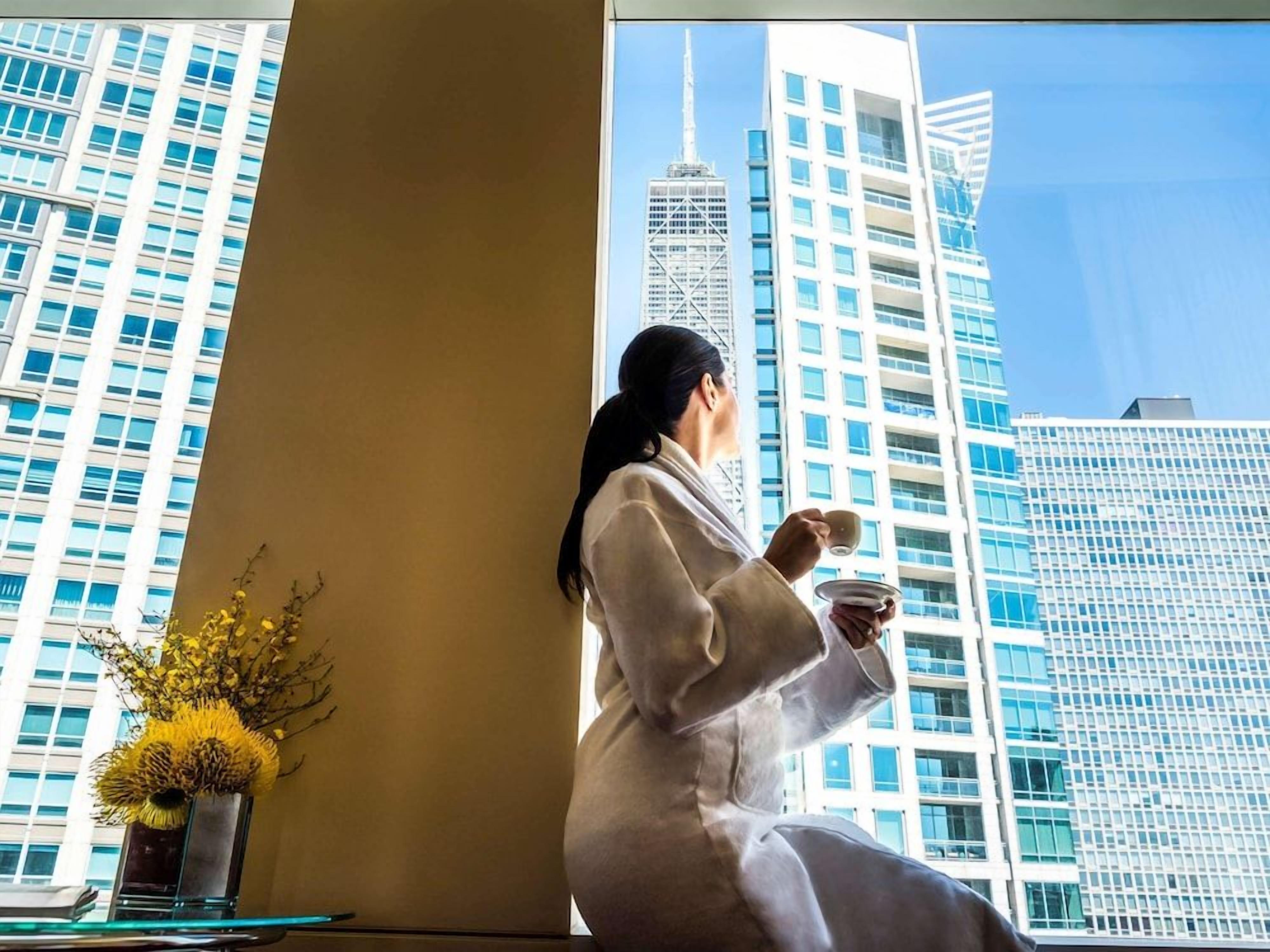 A person in a robe sitting by a window with a view of tall buildings.