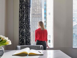 Une personne se tient près d’une fenêtre, d’où l’on aperçoit un paysage urbain. Sur une table, on voit un livre ouvert et, dans un vase, un bouquet de fleurs.