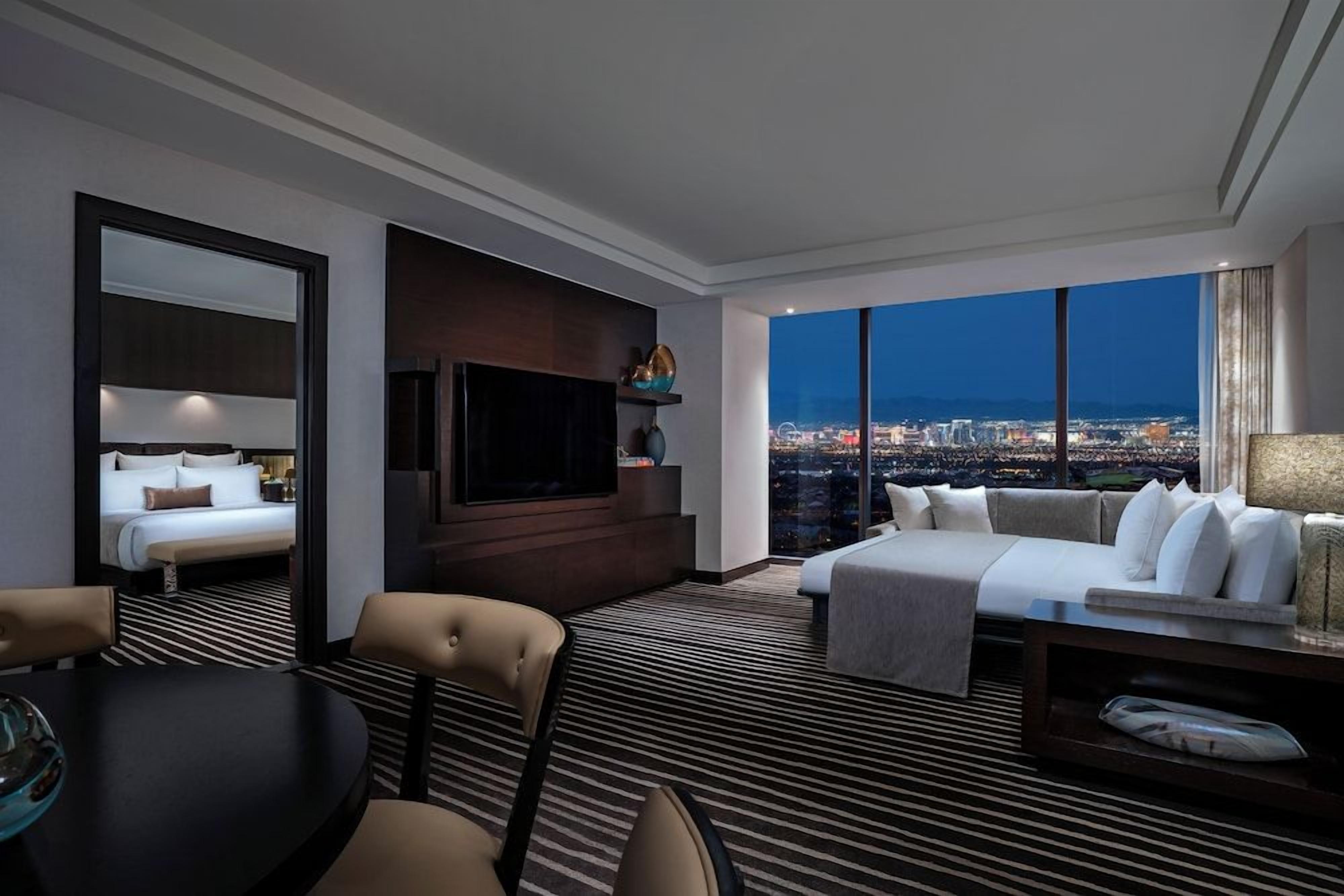 Quarto de hotel com cama, sofá, televisão, mesa de jantar e vista para a paisagem urbana.