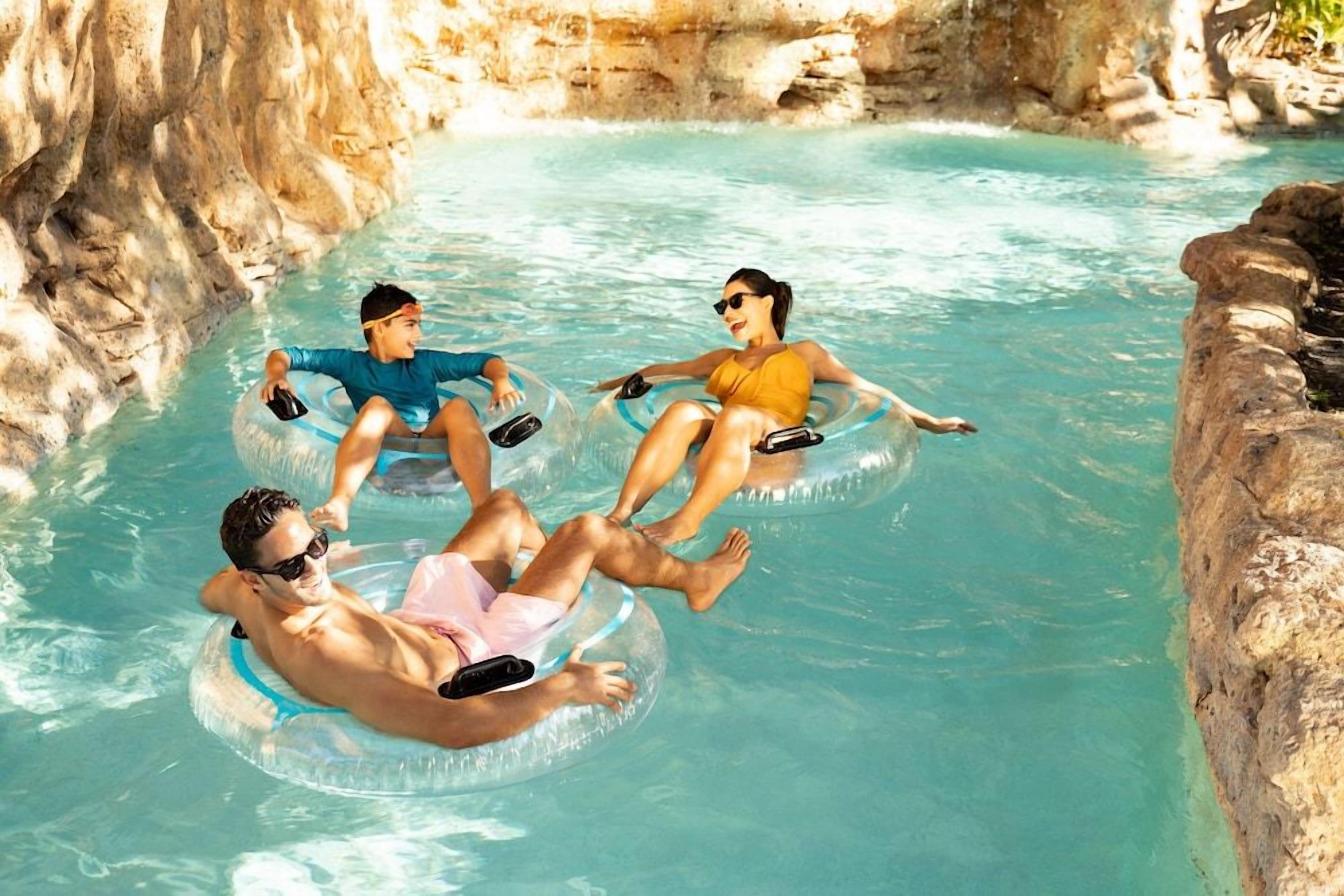 Three people floating on inflatable rings in a clear pool with rocky surroundings.