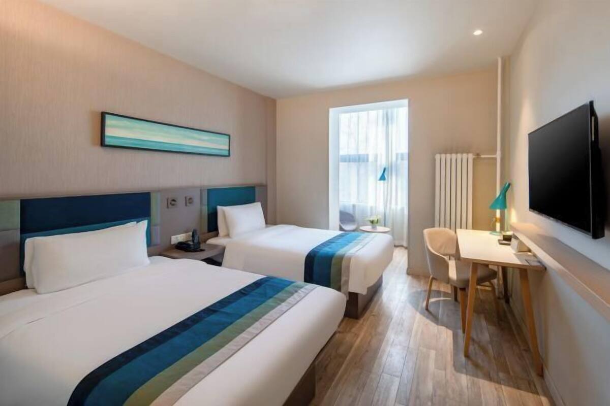 A hotel room with two beds, a desk, a chair, a TV, and a window with curtains.