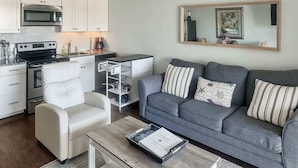 Ein kompakter Wohnraum mit Küchenzeile, einem grauen Sofa, einem weißen Sessel und einer Kaffeemaschine.