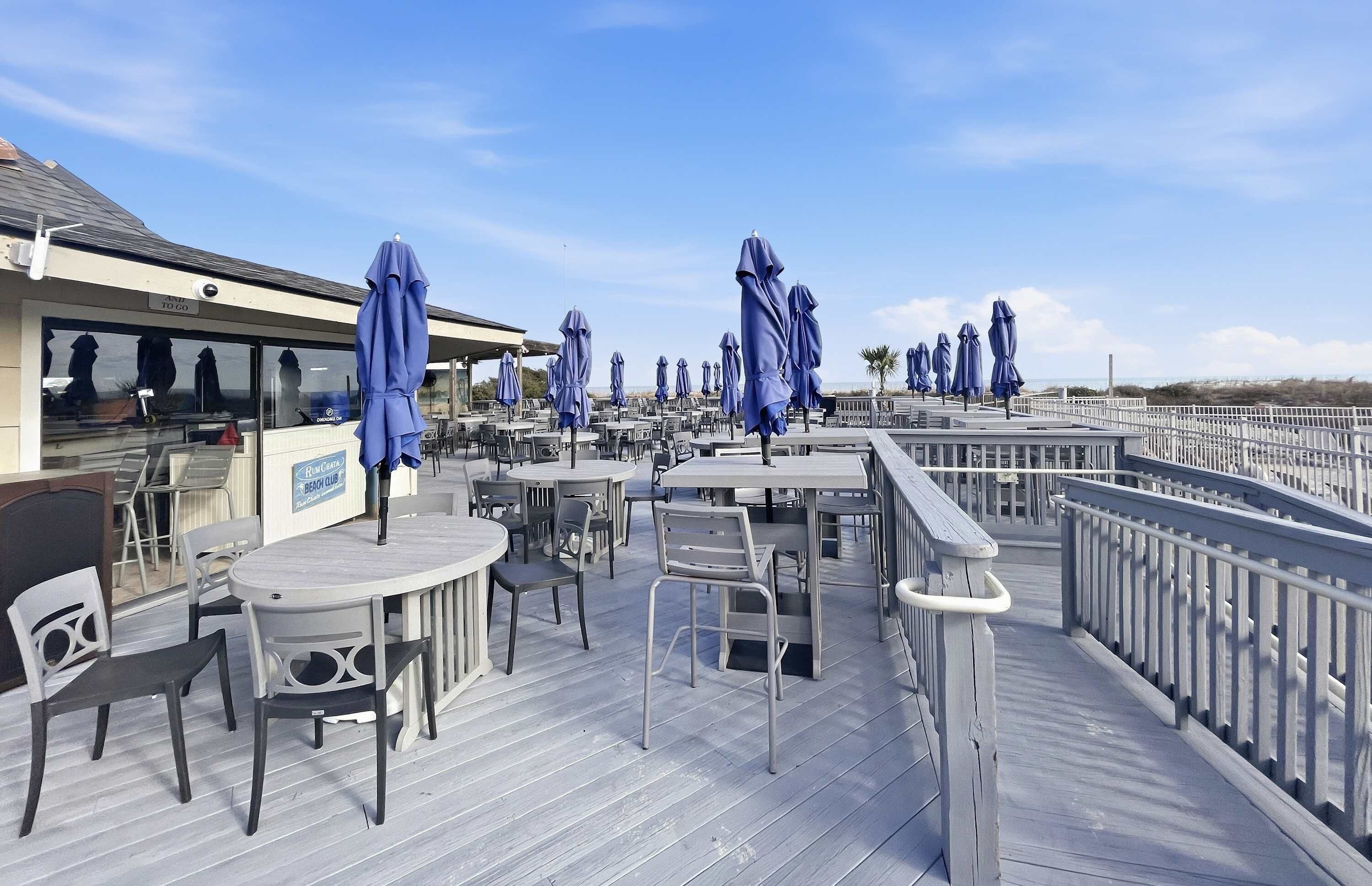 Une terrasse extérieure avec des tables et des chaises, des parasols bleus et un panneau indiquant l’emplacement d’un club de plage.