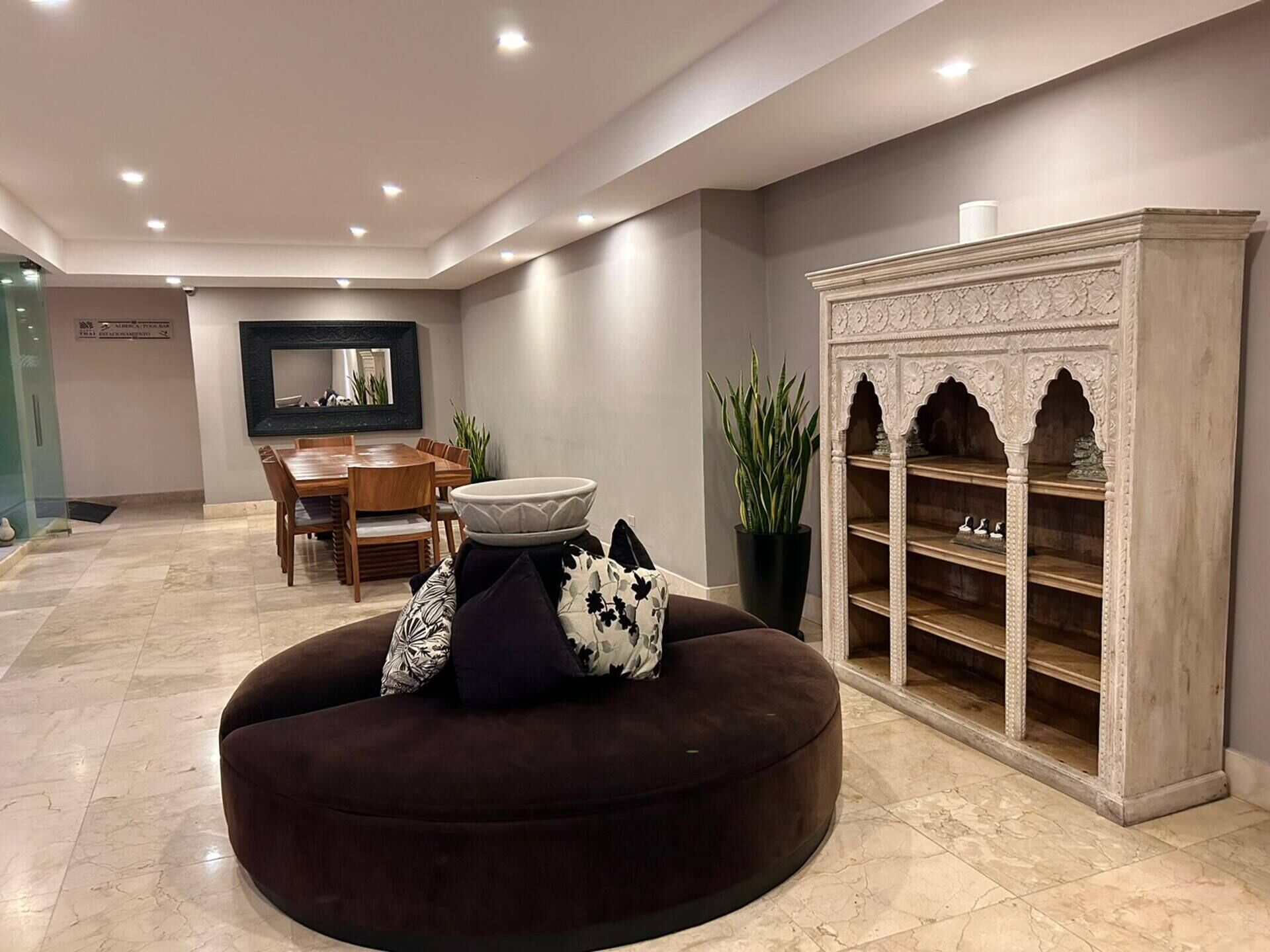 A modern living room with a dark round ottoman, a dining area with a wooden table and chairs, and a decorative cabinet with shelves.