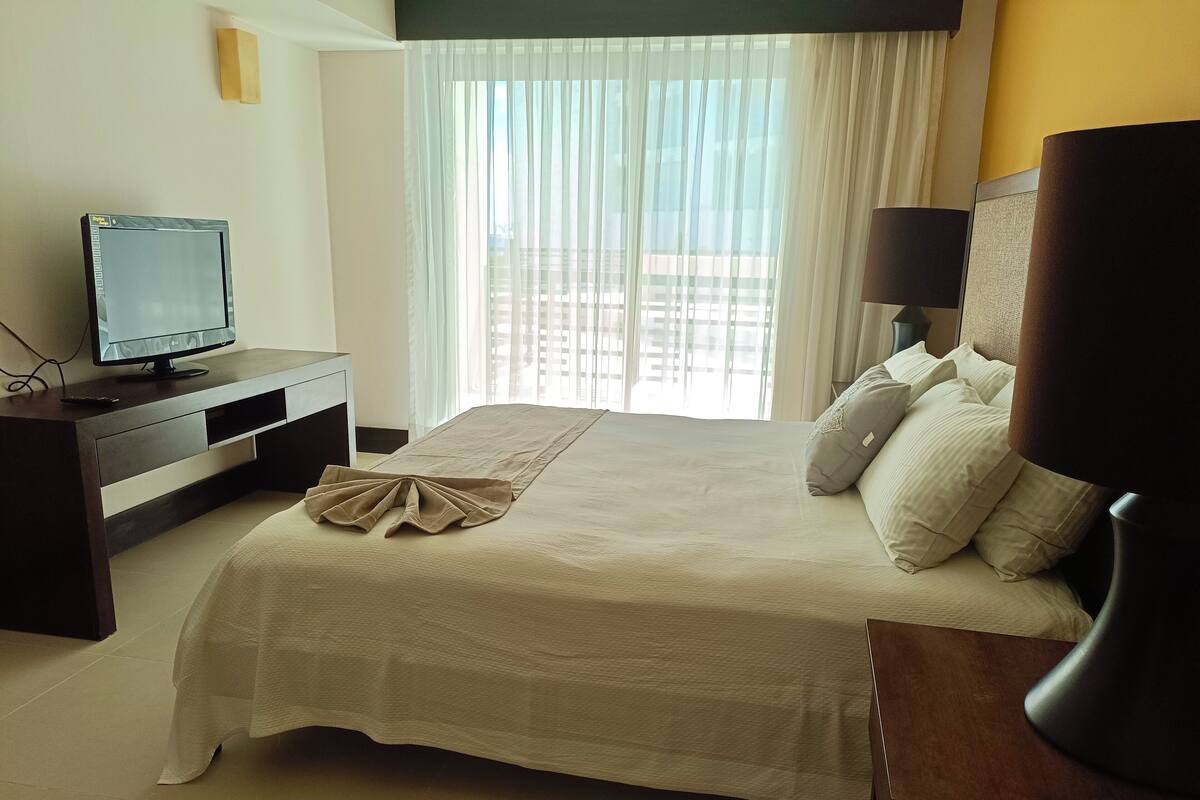 Hotellihuone, jossa on sänky, työpöytä tietokoneella, televisio ja ikkuna, jossa on verhot.