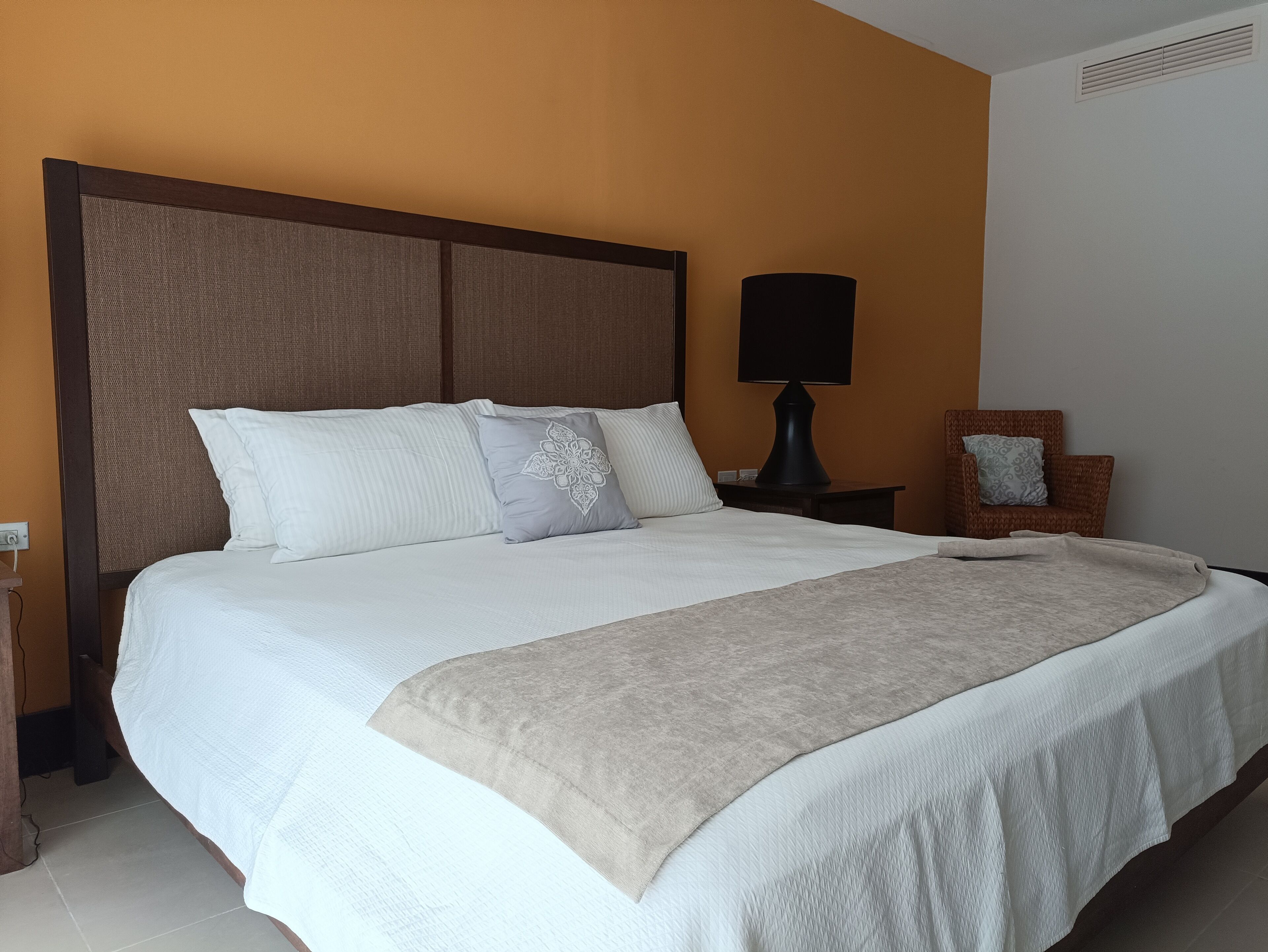 A neatly made bed with white linens and a beige throw blanket, a dark wooden headboard, a bedside lamp, and a wicker chair with a cushion.