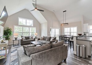 A spacious living room with a large gray sectional sofa, a coffee table, and a dining area with a bar. There are large windows providing natural light and a ceiling fan.
