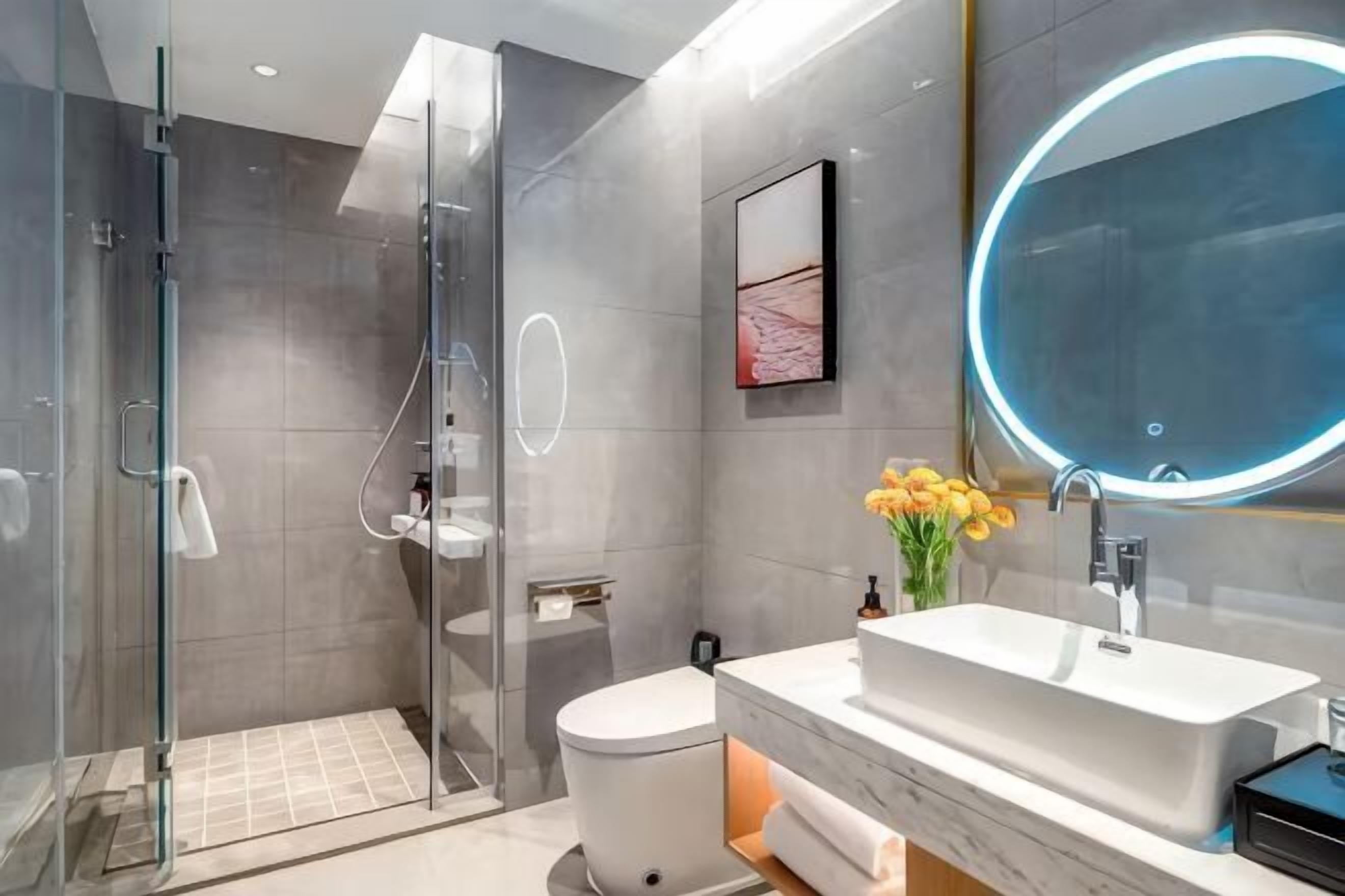 A modern bathroom with a glass shower enclosure, a white toilet, a sink with a marble countertop, and a round mirror with a light.