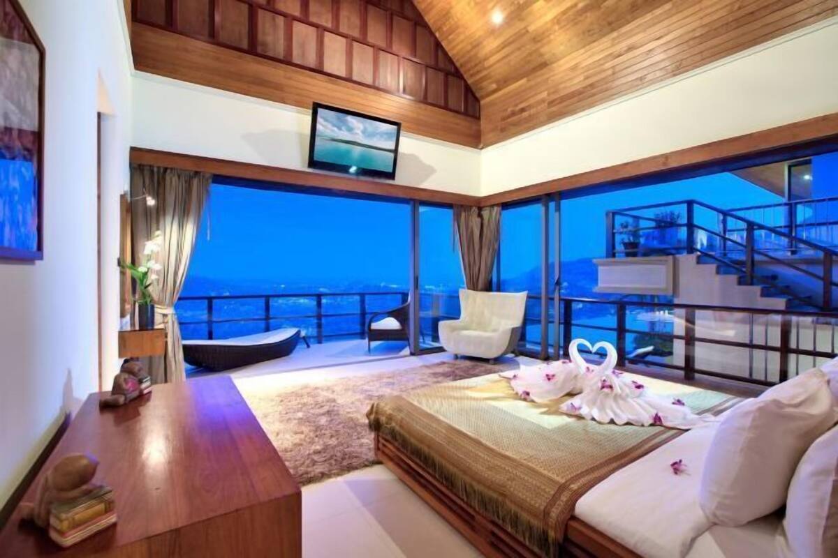 Een slaapkamer met een groot bed, een televisie, uitzicht op zee, een balkon en een trap.