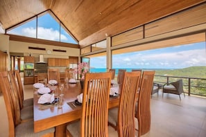 Un comedor con una mesa y sillas de madera, vista al océano y una cocina al fondo.