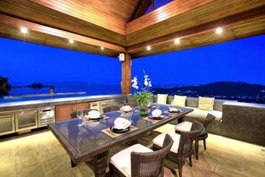 An outdoor dining area with a long table, chairs, and a view of the ocean at dusk. - Baan Grand Vista - Panoramic Sea View (Koh Samui)