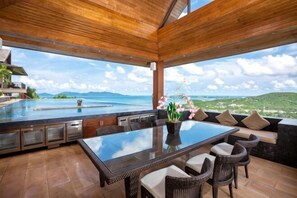 A spacious outdoor dining area with a glass table, wicker chairs, and a view of a mountainous landscape. - Baan Grand Vista - Panoramic Sea View (Koh Samui)