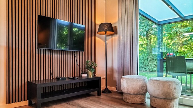 Een moderne woonkamer met een wand van houten latten, een zwarte entertainmentkast, een vloerlamp en twee ronde poefs.