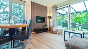 Een moderne woonkamer met een houten eettafel, zwarte stoelen, een aan een gestreepte muur bevestigde televisie en een glazen deur naar een terras.