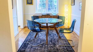 Un coin repas avec une table ronde en verre, quatre chaises bleues, un tapis à motifs, un lustre et une fenêtre donnant sur un paysage verdoyant.