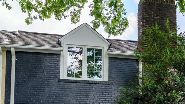 Une maison à deux étages avec une façade en pierre et un toit sombre. On y trouve une porte d’entrée avec un petit porche, et deux chaises bleues sont placées à l’extérieur.