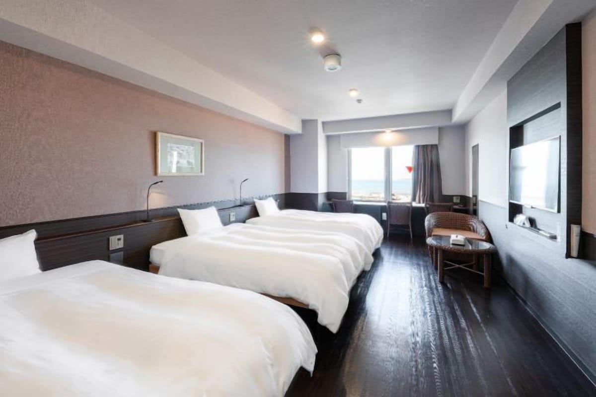 A hotel room with three beds, a small seating area, and a large window.