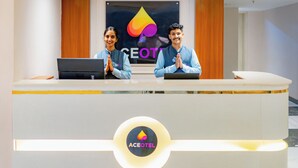 A reception desk with two staff members standing behind it, a logo on the wall, and a logo on the desk. - Aceotel Premier Vijay Nagar (Indore)