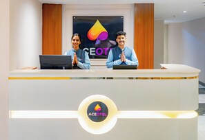 A reception desk with two staff members standing behind it, a logo on the wall, and a logo on the desk. - Aceotel Premier Vijay Nagar (Indore)