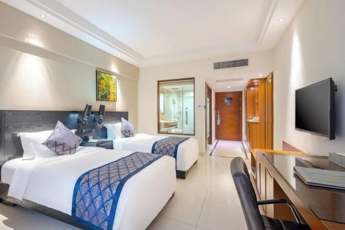Une chambre d’hôtel avec deux lits, un bureau, une chaise, une télévision et une salle de bain visible à travers une porte ouverte.