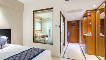 A hotel room with a bed, a large mirror, a bathroom visible through the mirror, and a wooden wardrobe.