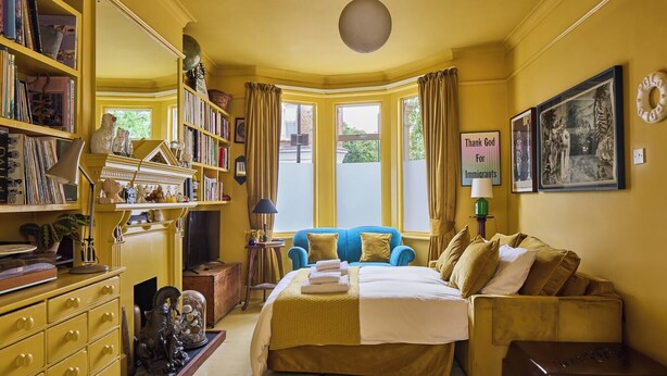 A room with a bed, a blue armchair, a bookshelf, and a window with curtains. - Charming East London Apartment (London)