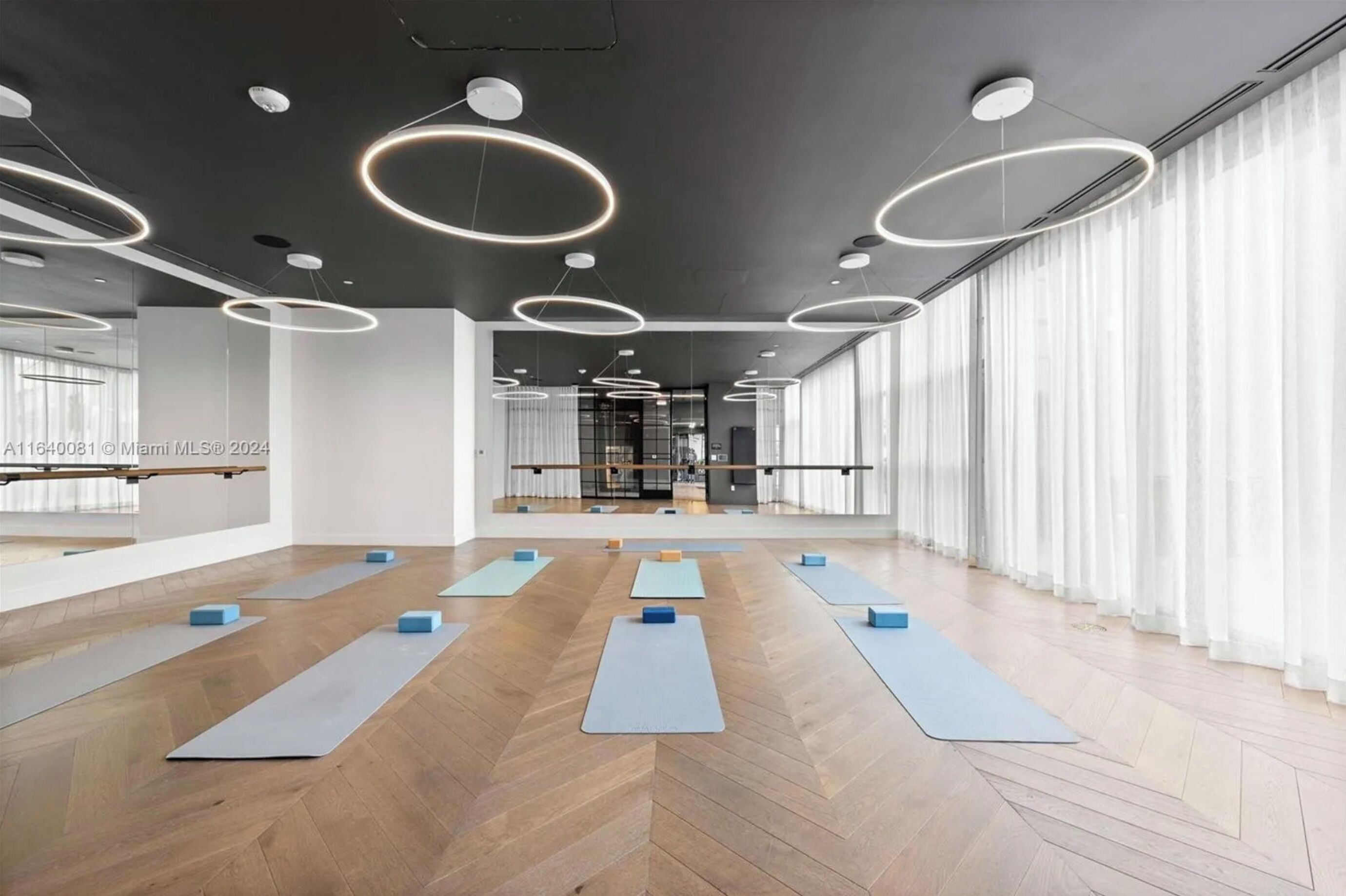 Une pièce spacieuse équipée de tapis et de blocs pour les exercices, de grandes fenêtres avec des rideaux légers et de luminaires circulaires modernes au plafond.