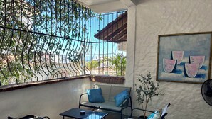Ein Balkon mit Metallgeländer, einem Glastisch, zwei Stühlen, einem blauen Kissen, einem Blumentopf und einem Bild von Wassermelonen.