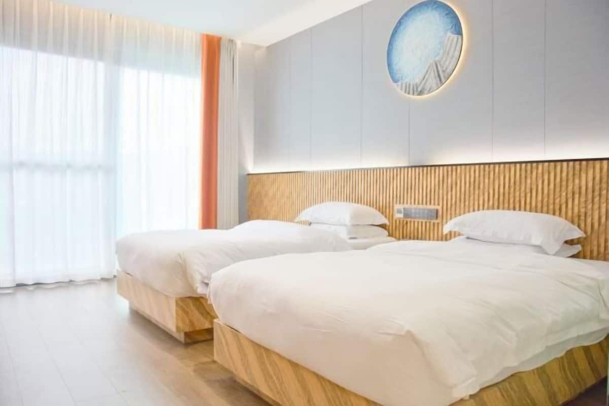 A hotel room with two beds, a wooden headboard, a wall-mounted light fixture, and a window with sheer curtains.