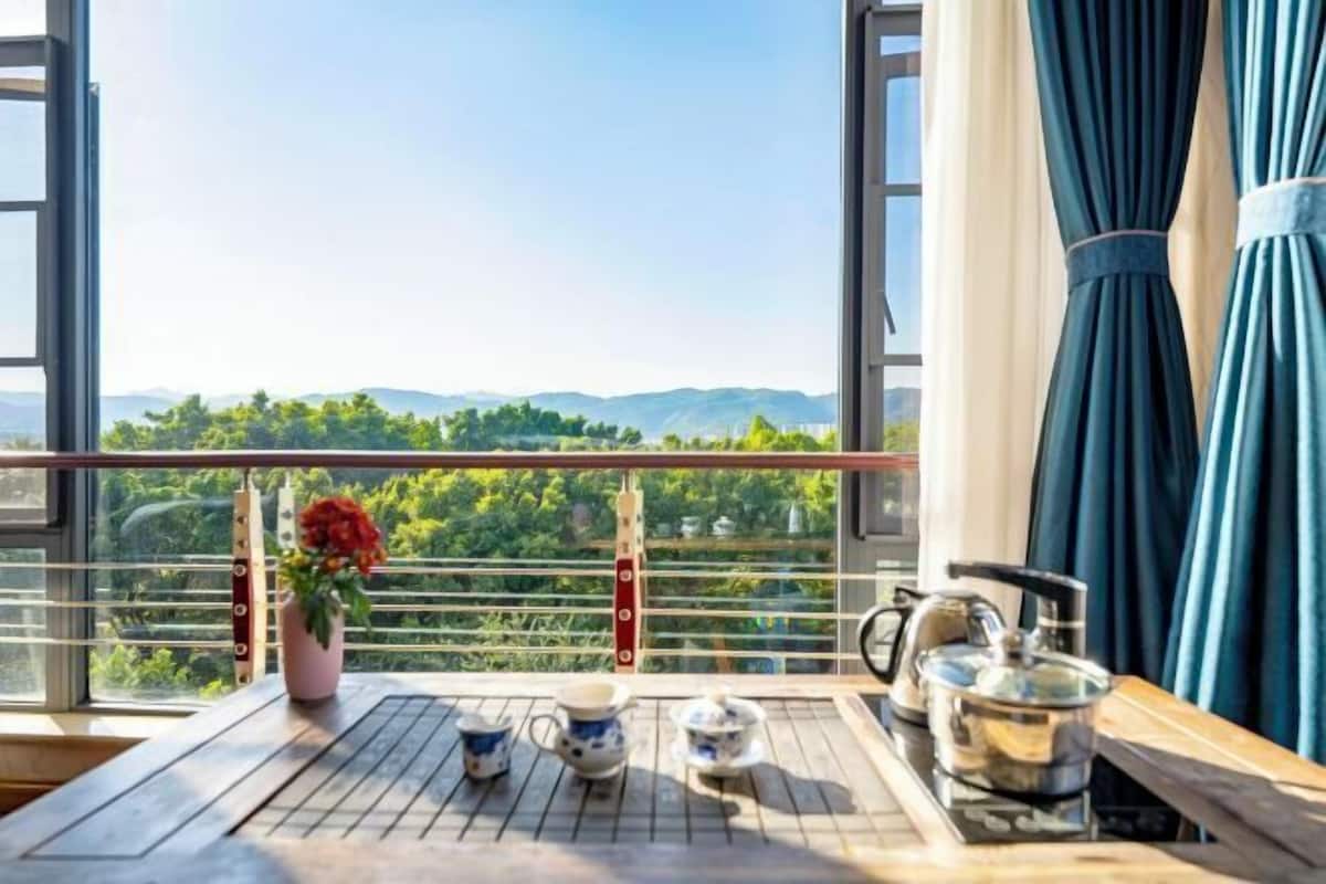 A balcony with a table set for tea, a flower vase, and a view of greenery and hills.