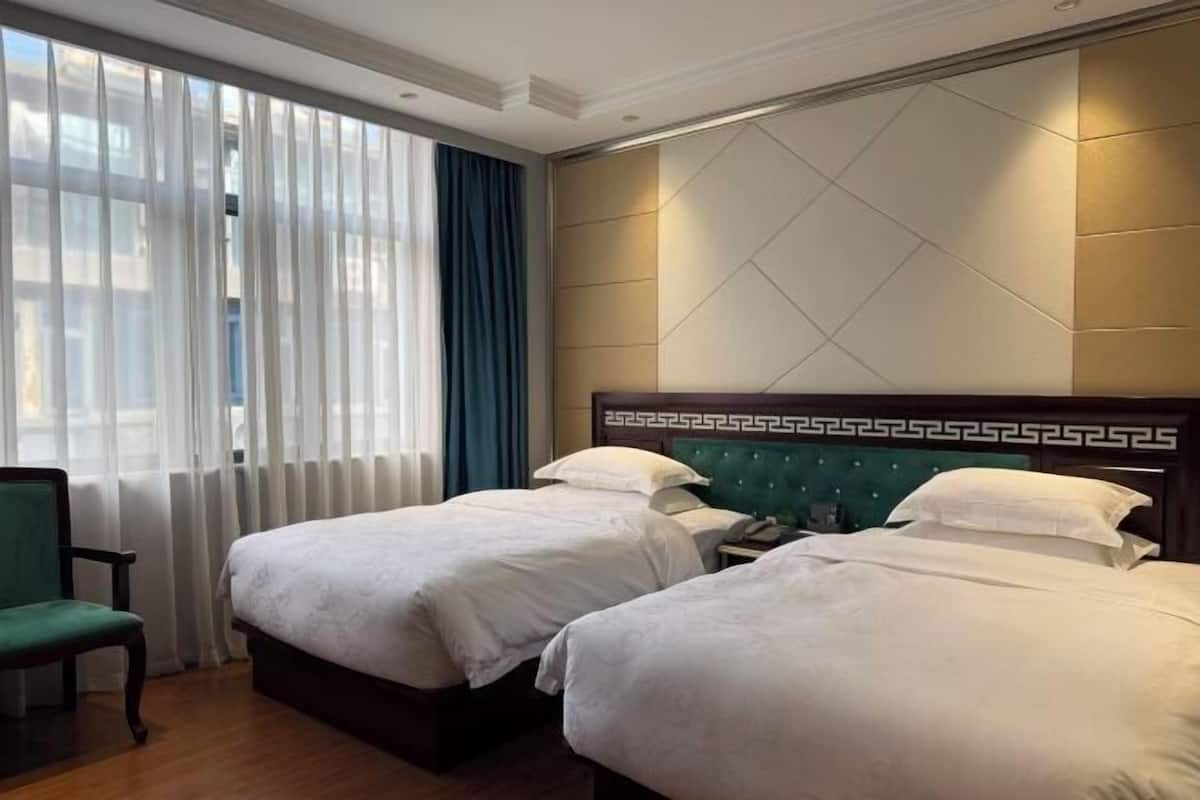 A hotel room with two beds, a green chair, a nightstand, and a window with curtains.